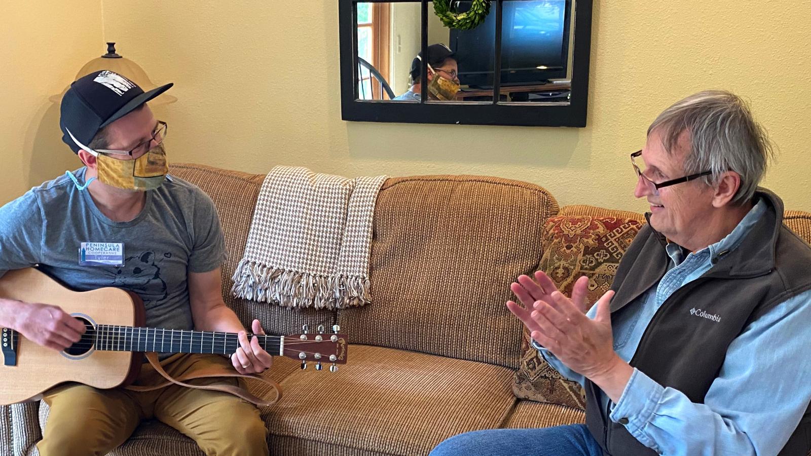 Photo of one person sitting on couch with guitar while other person listens.